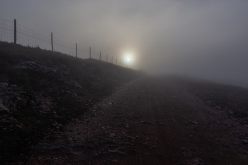 Low sun filtered through fog on a mountain, with a fence near a road