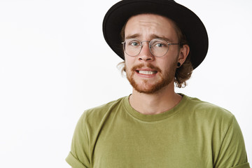 Yikes, I am in trouble. Portrait of worried sad cute guy with beard in glasses and hat frowning clenching teeth, feeling guilty as making bad thing to friend standing nervous over gray background