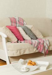 Beige sofa with plaid, colorful pillows (pink, grey, white) and coffee with croissantsin the living room.