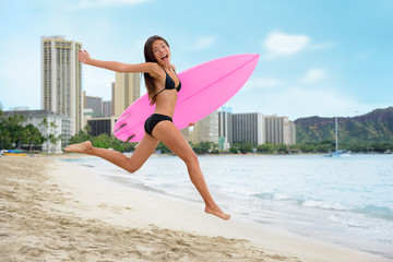 Surfing summer fun woman jumping of joy on beach with surfboard. Surf vacation lifestyle. Asian girl in bikini, sport and fitness active concept.