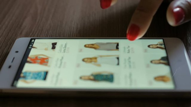 Woman looks at the goods  in the online clothing store. People using Smartphone, online shopping. Girl is choosing a shirt. Close-up. 4K UHD. Screen is blurred.