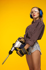 Beautiful young woman in goggles and headphones worth holding perforator drill isolated on yellow background