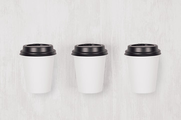 Coffee mockup - set of three white paper cups with black caps on white wood board, top view. Modern soft light elegant concept for branding identity, advertising and your text and design.