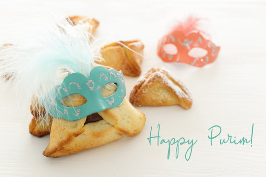 Purim celebration concept (jewish carnival holiday). Traditional hamantaschen cookies with cute mask over white wooden table.