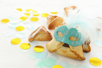 Purim celebration concept (jewish carnival holiday). Traditional hamantaschen cookies with cute mask over white wooden table.