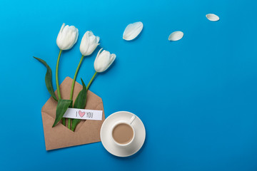 A bouquet of white tulips and a cup of coffee with a love note on a blue background. Top view with place for your congratulations.
