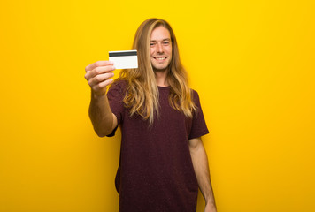 Blond man with long hair over yellow wall holding a credit card