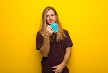 Blond man with long hair over yellow wall holding a hot cup of coffee