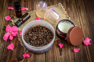Coffee body scrub, sugar and coconut oil, essential oils, massage vacuum jars on dark wooden rustic table with pink flowers. Homemade cosmetic for peeling and spa care.