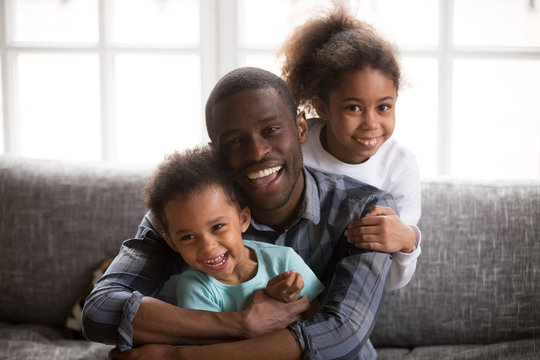 Happy african american dad and mixed race children, cheerful black family daddy embracing little son daughter sitting on sofa at home looking at camera, loving single parent with 2 cute kids portrait