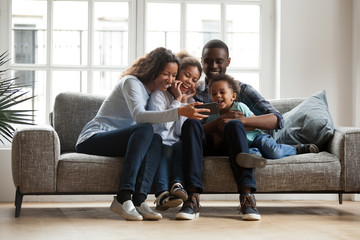 Happy african family with 2 children son daughter having fun with gadget on couch using smartphone at home, black parents and kids laugh watch funny video, make video call selfie on phone online app
