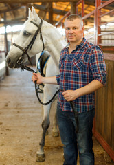 Positive man farmer  with white horse standing at stabling indoor