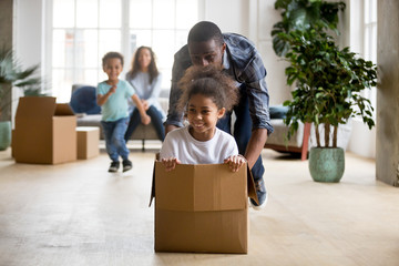 African dad playing with mixed race daughter riding in box on moving day concept, little kids enjoy...