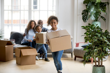 Happy mixed race kids running carrying boxes on moving day, black family tenants relocation concept, african american parents and children laughing playing in new home flat having fun in living room