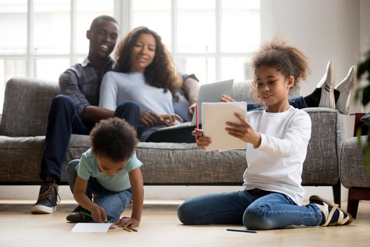 Mixed race children playing drawing with colored pencils on warm floor while happy african parents relax on couch in living room, cute kids siblings having fun at home, black family leisure time