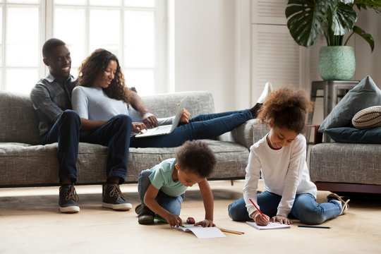Black family relaxing at modern home, african american parents resting on sofa with laptop while mixed race kids playing together drawing on warm floor, children have fun with mom dad in living room