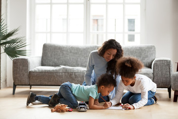 Loving black mom and little mixed race children drawing with colored pencils in living room, happy single african mum helping kids with creative activity at home, underfloor heating, family hobbies