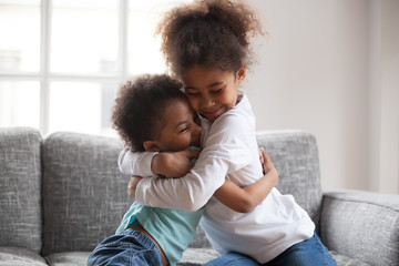 Cute happy african american siblings hugging cuddling feeling love and connection, smiling mixed race kid girl sister embracing little boy brother sitting on couch, 2 children good relationships - Powered by Adobe