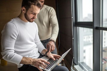 Two concentrated black and white businessmen in casual wear discussing project sitting at office desk, near the window, using pc laptop, looking at screen, make presentation, fulfill task.