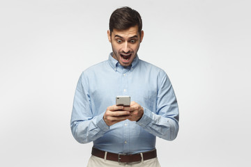Young business man looking at his phone with surprise expression, isolated on gray background