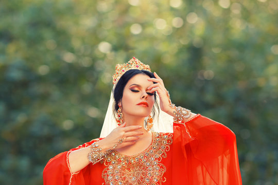 charming delightful Turkish lady in bright red scarlet light dress puts her hands to a gorgeous face, mistress Roksolana with amazing expensive luxury crown on dark hair, gold jewelry and ornaments