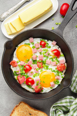 Fried eggs in black pan with ham, cherry tomatoes and spring green onions. Delicious Breakfast with toast and butter. The classic English lunch