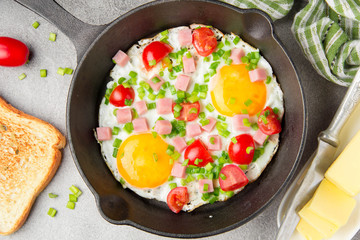 Fried eggs in black pan with ham, cherry tomatoes and spring green onions. Delicious Breakfast with toast and butter. The classic English lunch