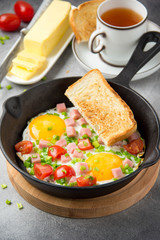 Fried eggs in black pan with ham, cherry tomatoes and spring green onions. Delicious Breakfast with toast and butter. The classic English lunch