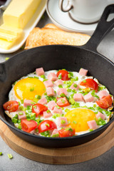 Fried eggs in black pan with ham, cherry tomatoes and spring green onions. Delicious Breakfast with toast and butter. The classic English lunch