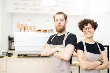 Positive confident young team of coffee shop staff standing at espresso machine and looking at camera while crossing arms on chest