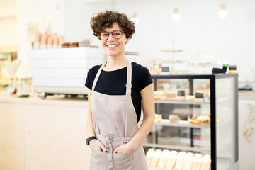 Cheerful confident beautiful lady barista with curly hair standing in bakers store and looking at camera while holding hands in apron pockets