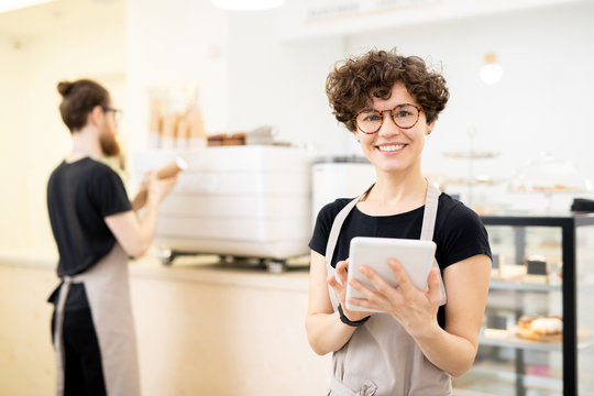 Smiling pretty waitress in apron and glasses standing in coffee shop and looking at camera, she using tablet to take order