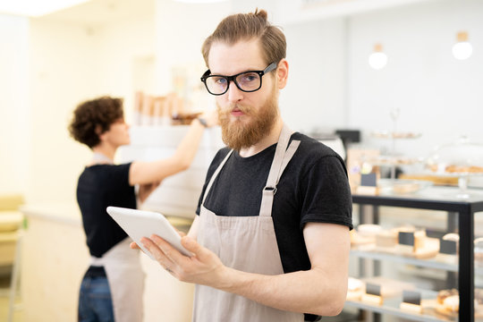 Serious handsome hipster young bearded waiter in glasses standing in bakery store and looking at camera while using digital tablet
