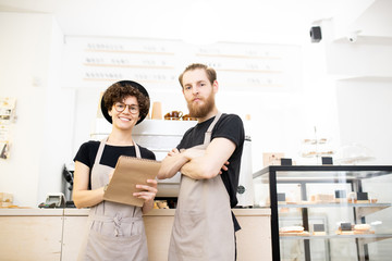 Fototapeta premium Content confident team of coffee shop: smiling young man and woman in aprons standing at counter and looking at camera while analyzing notes in sketchpad