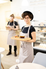 Content attractive hipster lady waitress in apron carrying wooden tray with coffee cup and looking at camera in coffee shop