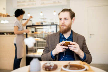 Serious thoughtful bearded man in earphones sitting at table and drinking coffee while relaxing in coffee shop
