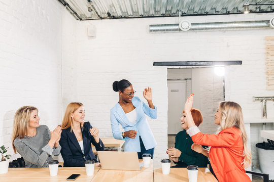 Cheerful young dark-skinned African female Chief giving high five with her caucasian co-workers, looking at each other with happy smiles, cheering and celebrating successful teamwork at meeting.