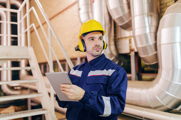 Young Caucasian man in protective suit using tablet while standing in heating plant.