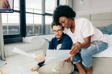 Young multiracial couple sit on floor in spacious apartment, checking bills, study documents, prepare financial report Family relation and common deals concept