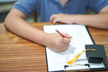 Asian children young kid write answer in a quiz examination test study learn with graduation gap hat on wooden table in school : bright genius education concept.