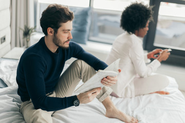 Fototapeta premium Young beautiful interracial couple in bed where everyone is busy with his own things, not paying attention to his partner