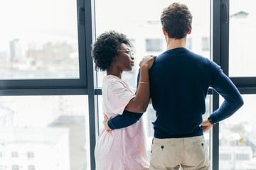 Backside view of young mixed race lovers staying in hotel, Hispanic man hugging African Woman while resting in modern apartment with big window interior