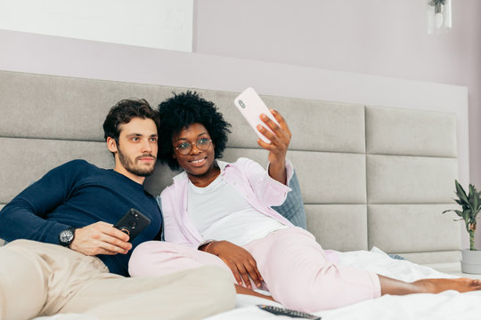 Cheerful young mixed race loving couple lying in bed and making selfie on light gray background - Powered by Adobe