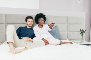 Mixed race couple resting on bed and watching TV together in day-off, enjoying togetherness and domestic atmosphere