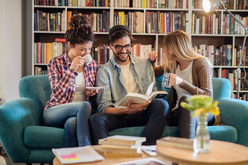 Three smiling students sitting on sofa in library and looking at book.