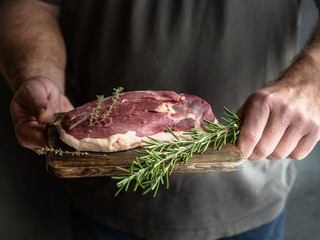 Man's hands holding raw uncooked duck breast with rosemary and thyme on wooden cutting board.