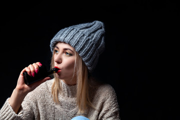 Blonde in a knitted sweater and hat holds an electronic cigarette vape in her hand with a manicure...