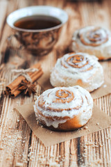 Freshly baked warm iced cinnamon buns on baked paper. Sweet homemade pastry baking on with cup of coffee and spices on wooden background.