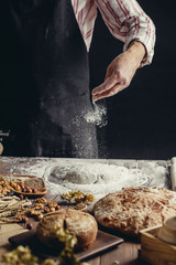 Man in black apron sprinkling some flour on dough isolated over dark studio background. Hands kneading dough, cropped view. People and baking concept