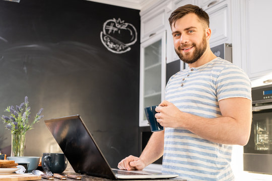 Waist up portrait of handsome bearded man using laptop in kitchen while standing at counter and drinking coffee  looking at camera, copy space